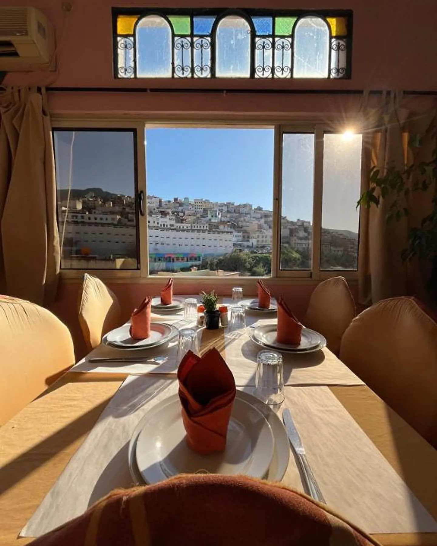 Une table dressée face à la fenêtre, la ville blanche de Moulay Idriss au-delà, sous un vitrail en arc.